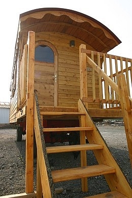 Escalier de la roulotte et sa terrasse en bois, la porte d'entrée est cintrée avec le carreaux en demi-lune.